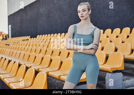 Hübsche und blonde Sportlerin in Aktivkleidung posiert mit überkreuzten Armen neben gelben Stadionstühlen Stockfoto