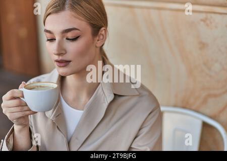 Elegante blonde Frau im stilvollen Trenchcoat hält eine Tasse Kaffee, während sie im Café sitzt, Cappuccino Stockfoto