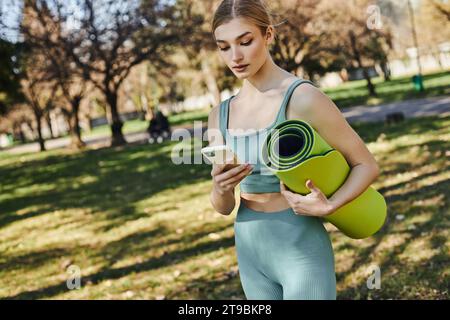 Hübsche Sportlerin in aktiver Kleidung mit Smartphone und Fitnessmatte im Park Stockfoto