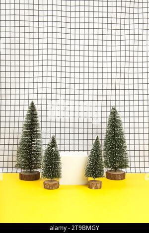 Ein weißer Holzwürfel, umgeben von mehreren Miniatur-weihnachtsbäumen auf einem schwarz-weißen Gitterhintergrund. Leerer Objektständer. Minimales still li Stockfoto