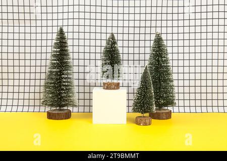 Ein weißer Holzwürfel, umgeben von mehreren Miniatur-weihnachtsbäumen auf einem schwarz-weißen Gitterhintergrund. Leerer Objektständer. Minimales still li Stockfoto