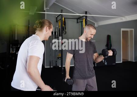 Mann, der mit Hantel trainiert, mit Trainer, der seinen Fortschritt überprüft Stockfoto