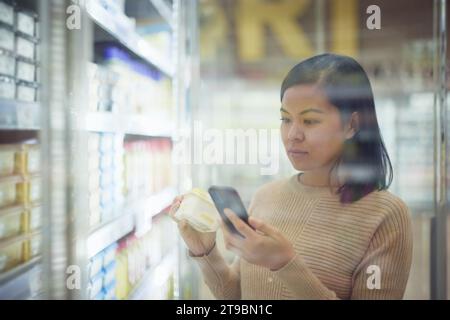 Frau im Supermarkt, die vor dem Kühlschrank steht und das Handy benutzt Stockfoto