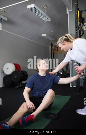 Lächelnde Frau, die dem Mann, der auf einer Trainingsmatte im Fitnessclub sitzt, eine Flasche Wasser gibt Stockfoto