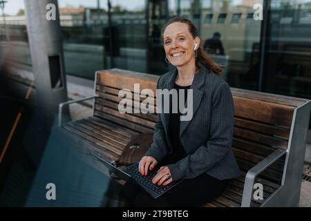 Porträt einer glücklichen Geschäftsfrau, die mit Laptop auf der Bank sitzt, während sie am Bahnhof wartet Stockfoto