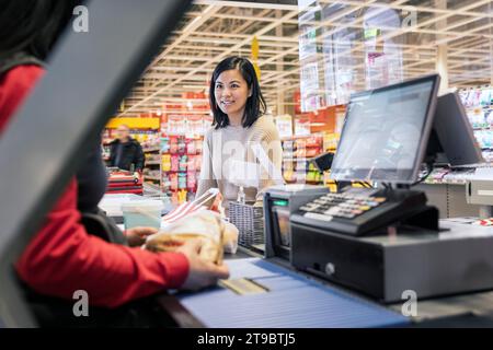 Lächelnde junge Kundin, die sich beim Checkout im Supermarkt mit der Kassiererin unterhalten hat Stockfoto