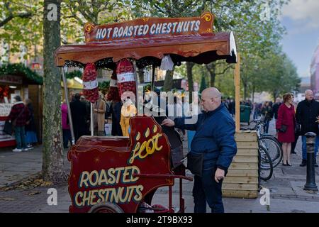 Ein festlicher rot-brauner Wagen mit einem Mann, der heisse geröstete Kastanien auf einem belebten Weihnachtsmarkt in York, Yorkshire, Großbritannien zubereitet. Stockfoto