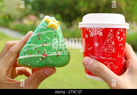 Hände halten Weihnachtsbaum geformten Donut und Feiertagsthema Take-away Coffee Cup im Garten Stockfoto