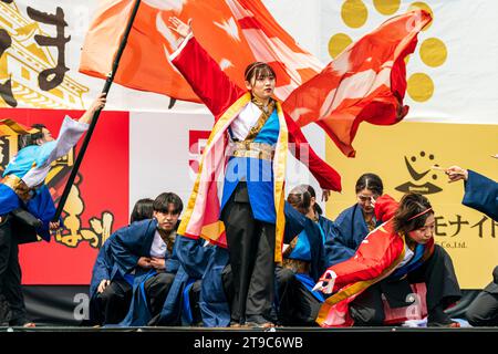 Nahaufnahme einer jungen japanischen Yosakoi-Tänzerin, die mit den Armen zur Seite gestreckt steht, ein Banner hinter ihr wirbelt und andere Tänzerinnen hocken. Stockfoto