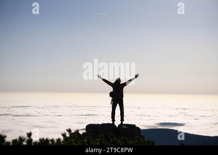 Ein Mann mit ausgestreckten Armen in den tschechischen Bergen, der über Wolken steht, die den Boden darunter verdecken und einen surrealen Blick schafft. Freiheit. Stockfoto