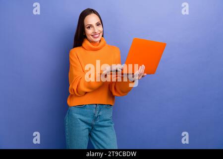 Foto von reifer Geschäftsfrau in orangem gestricktem trendigen Pullover, der macbook pro Modell für die Arbeit isoliert auf violettem Hintergrund hält Stockfoto