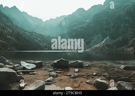 Wo Ruhe fließt – die sanfte Umschlingung eines Flusses aus Steinen, eingerahmt von majestätischen Bergen am fernen Horizont. Ein sanfter Nebel liegt über dem Berg Stockfoto