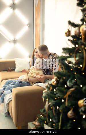 Eine Familie sitzt auf dem Sofa neben einem wunderschön dekorierten Neujahrsbaum. Dad, Mom und kleiner Sohn im Wohnzimmer, das Weihnachten dekoriert ist. Weihnachten. Stockfoto