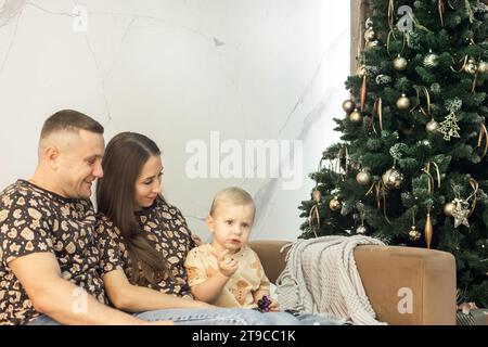 Eine Familie sitzt auf dem Sofa neben einem wunderschön dekorierten Neujahrsbaum. Dad, Mom und kleiner Sohn im Wohnzimmer, das Weihnachten dekoriert ist. Weihnachten. Stockfoto