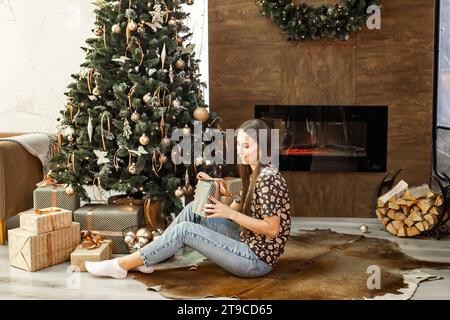 Ein schönes Mädchen dekoriert einen Weihnachtsbaum in einem großen Wohnzimmer mit Kamin. Wunderschöne Innenausstattung. Konzept des Wartens auf den Urlaub. Stockfoto