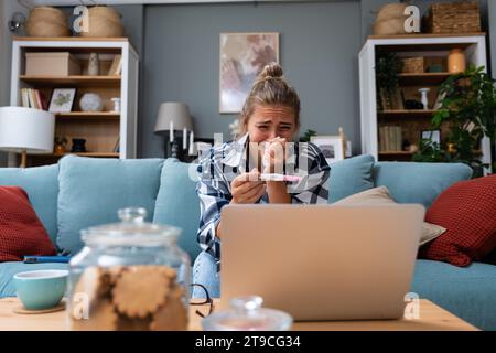 Einzelne traurige Frau, die sich über einen Schwangerschaftstest beschwert, sitzend auf einer Couch im Wohnzimmer zu Hause, gestresste Frau, positive oder negative medizinische Taten Stockfoto