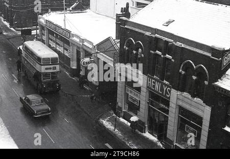 1960er Jahre, historisch, winterlich und ein Blick über eine Straße in Oldham, England, Großbritannien, mit schneebedeckten Dächern und unten zeigt die Garage und den Ausstellungsraum von Henleys und auf der Straße einen Bus und ein Auto der damaligen Zeit. Stockfoto