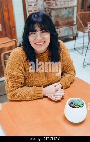 Vertikales Bild der jungen lateinamerikanischen venezolanischen Frau, in braun gekleidet, sitzt mit den Händen auf dem Tisch und sieht die Kamera lächelnd und glücklich im Kaffee wartend an Stockfoto