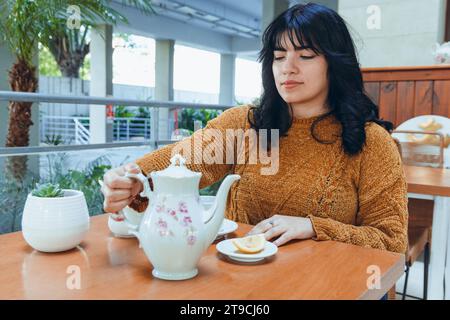 Die junge glückliche lateinfrau sitzt allein im Restaurant mit einer Teekanne und gießt sich während des Sonnenuntergangs Tee in Porzellantasse Stockfoto