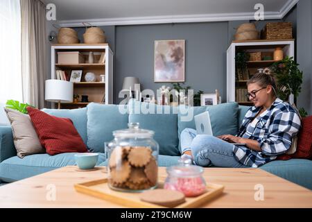 Junge Freiberuflerin in lässiger Kleidung, die auf einer bequemen Couch sitzt und ferngesteuert auf einem Laptop arbeitet. Eine Frau, die Urheberrechtschreiber oder Webentwickler ist, arbeitet bei Stockfoto