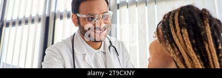 Konzentrieren Sie sich auf den fröhlichen indischen Arzt, der mit seinem verschwommenen afroamerikanischen Patienten Hand schüttelt, Banner Stockfoto