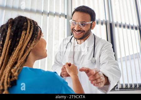 Konzentrieren Sie sich auf den fröhlichen indischen Arzt in Brille, der mit seinem verschwommenen afroamerikanischen Patienten die Hand schüttelt Stockfoto