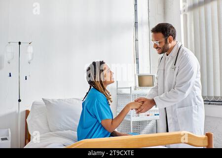 Fröhlicher indischer Arzt in Brille, der mit seinem fröhlichen afroamerikanischen Patienten die Hände schüttelt, Gesundheitswesen Stockfoto