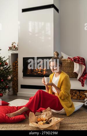 Happy Woman sitzt neben einem glühenden Kamin in einem gemütlichen Wohnzimmer, geschmückt mit einem Weihnachtsbaum und festlichen Dekorationen, und genießt eine Tasse heißes Getränk. Stockfoto