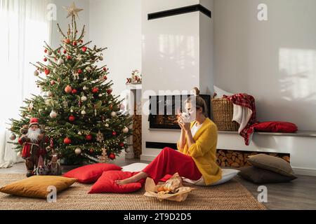 Happy Woman sitzt neben einem glühenden Kamin in einem gemütlichen Wohnzimmer, geschmückt mit einem Weihnachtsbaum und festlichen Dekorationen, und genießt eine Tasse heißes Getränk. Stockfoto