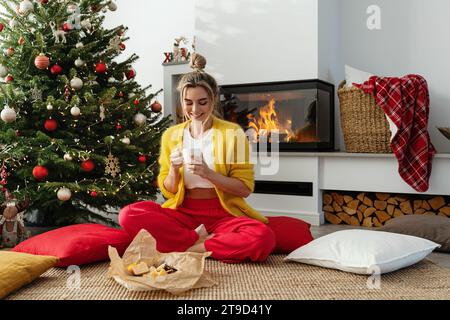 Happy Woman sitzt neben einem glühenden Kamin in einem gemütlichen Wohnzimmer, geschmückt mit einem Weihnachtsbaum und festlichen Dekorationen, und genießt eine Tasse heißes Getränk. Stockfoto