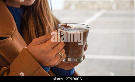 Eine Person, die heiße Schokolade im Café trinkt. Nahaufnahme weiblicher Hände, die einen Becher halten Stockfoto