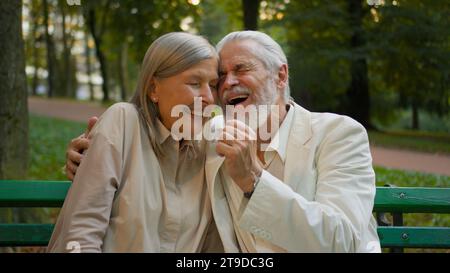 Nahaufnahme Porträt zwei ältere Rentner altes kaukasisches Paar sitzend Bank lachend Witze redend kuschelig umarmt Stadtpark draußen. Erholung Relaxwochenende Stockfoto