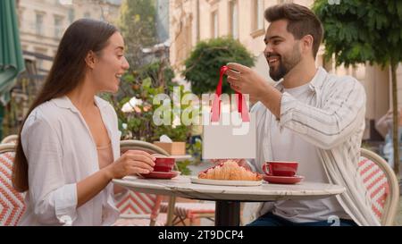 Glückliches europäisches Paar Mann gibt Frau ein Geschenk Überraschung Jubiläum Feier Emotionen Stadt draußen Café zusammen zarte Liebe Gefühle lächelnd Stockfoto