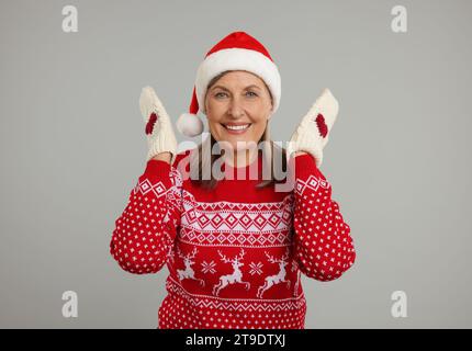 Glückliche Seniorin in Weihnachtspullover, Weihnachtsmütze und gestrickten Fäustlingen auf grauem Hintergrund Stockfoto