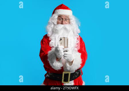 Frohe Weihnachten. Weihnachtsmann mit Smartphone auf hellblauem Hintergrund Stockfoto