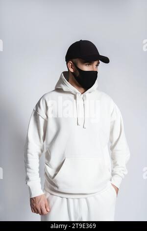 Junger Mann mit weißem Hoodie, schwarzer Baseballmütze und Stoffmaske vor grauem Hintergrund Stockfoto