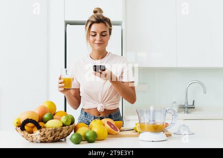 Junge Frau trinkt frisch gepressten, selbstgemachten Orangensaft und macht Fotos mit ihrem Smartphone Stockfoto