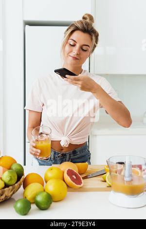 Junge Frau trinkt frisch gepressten, selbstgemachten Orangensaft und macht Fotos mit ihrem Smartphone Stockfoto