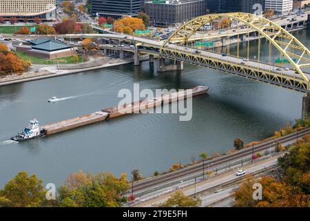 Die Farben des Herbstes steigen auf, wenn das Schleppboot Tracy Fedkoe leere Binnenschiffe den Monongahela River hinauf und unter die Fort Pitt Bridge in Pittsburgh schiebt, 27. Oktober 2023. Das U.S. Army Corps of Engineers Pittsburgh District verwaltet die oberen 127 Meilen des Ohio River und die Einzugsgebiete des Allegheny River und des Monongahela River, die an der Stelle in Pittsburgh zum Ohio River münden. Die drei Flüsse, die den Hafen von Pittsburgh bilden, werden für den Transport von Rohstoffen, Schüttgütern und Fertigwaren für viele Industrien in der Region genutzt. Es ist der zweitgrößte Binnenhafen und der zweitgrößte Hafen überhaupt Stockfoto