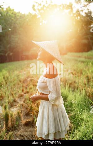 Glückliche lächelnde Frau mit Naturkleidung und asiatischem konischem Hut auf dem Reisfeld während des Sonnenuntergangs Stockfoto