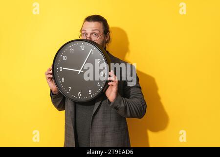 Junger Geschäftsmann, der hinter einer großen Uhr auf gelbem Hintergrund rausblickt Stockfoto