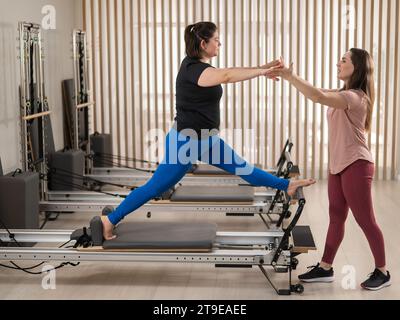 Übergewichtige Kaukasierin, die Pilates-Übungen auf Reformer mit Personal Trainer macht. Stockfoto