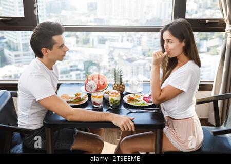 Glückliches Paar, das gesundes Frühstück in einem modernen Apartment mit großen Fenstern und Blick auf die Stadt isst Stockfoto