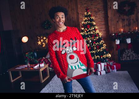 Foto von aufgeregtem funky Guy trägt roten hässlichen Weihnachtspullover, der weihnachtsoutfit im Appartementzimmer zeigt Stockfoto