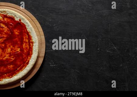 Pizzateig mit Tomatensauce auf einem runden Holzständer, auf dunklem Hintergrund, Draufsicht, Platz zum Kopieren von Text. Stockfoto