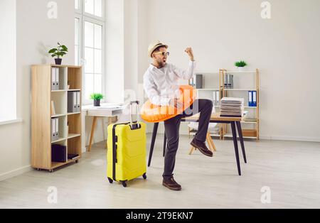 Glücklicher Geschäftsmann mit Koffer und Strandring geht in den Urlaub und hat Spaß im Büro Stockfoto