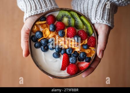 Frau, die gesundes Frühstück mit Früchten in Kokosnussschale hält. Joghurt mit Cornflakes und Heidelbeere, Erdbeere, Kiwi und Himbeere. Vegetarische Speisen Stockfoto