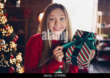 Foto von nettem netten guten Laune Schulmädchen mit blondem Haar tragen roten Pullover auspacken Geschenk am weihnachtsmorgen zu Hause drinnen Stockfoto