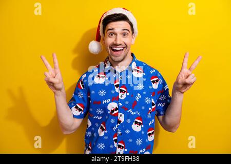 Holly fröhliche Weihnachtsfeier zwei Hände V Zeichen rot Hut Pom blau Hemd traditionelles Bild isoliert auf gelbem Hintergrund Stockfoto