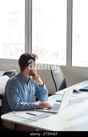 Geschäftiger lateinamerikanischer Geschäftsmann, der im Büro arbeitet, mit einem Laptop, der in der vertikalen Position wegblickt. Stockfoto